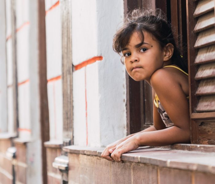 LA HABANA, CUBA - NOVEMBER, 6 , 2018: Thinking little girl hanging out of window of building with white facade in Cuba